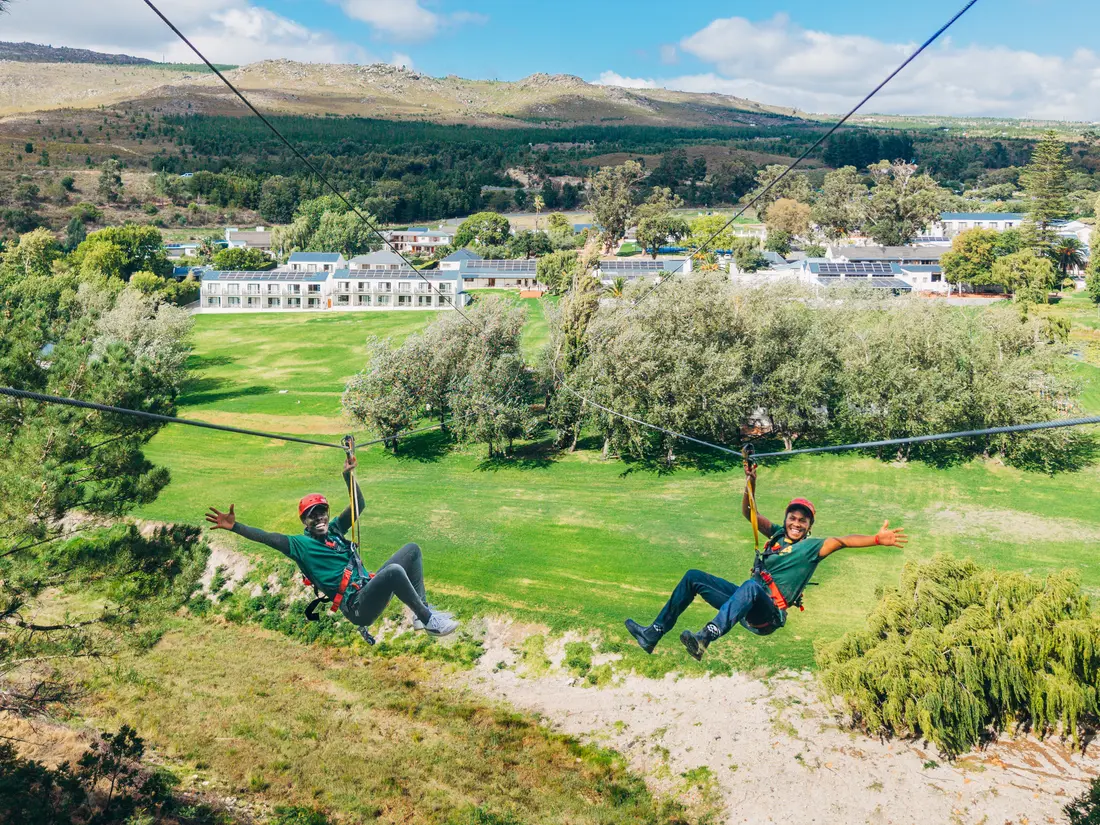 Two riders racing side by side on the dual zipline at Houw Hoek Zipline, with Houw Hoek Hotel and the Overberg valley below